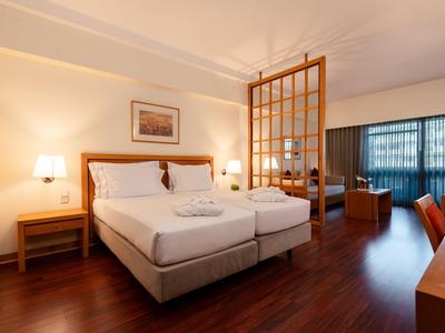 Camera d'hotel luminosa con letto matrimoniale, pavimento in legno e area salotto dietro un divisorio in legno.