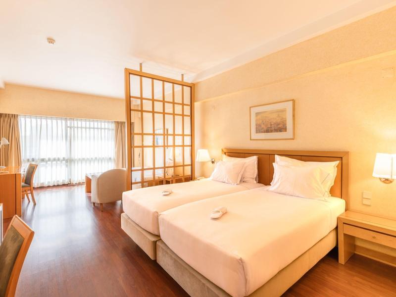 Camera d'hotel luminosa con due letti singoli, pavimento in legno e divisorio davanti alla finestra.