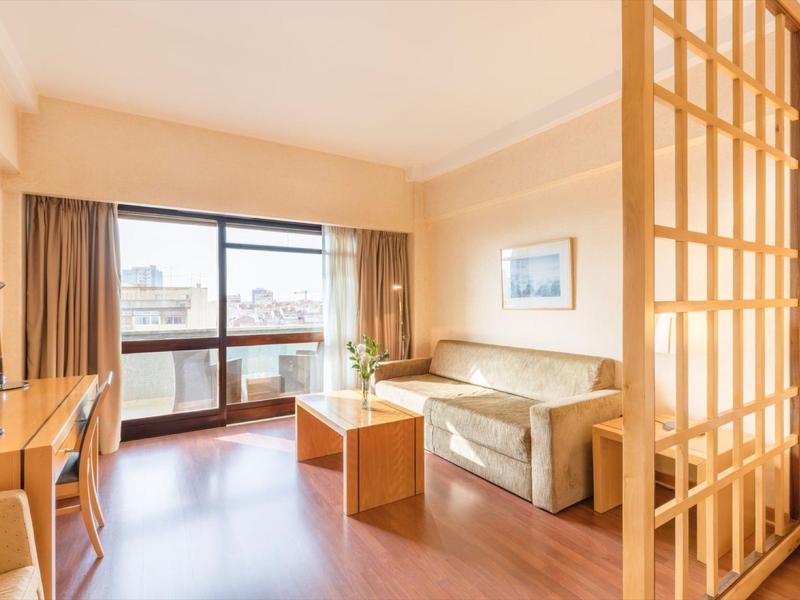 Camera d'hotel luminosa con divano, scrivania e balcone con vista sulla città.