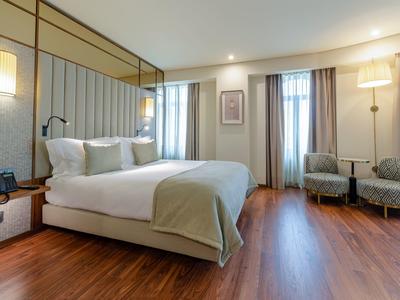 Camera d'hotel luminosa con letto grande, due poltrone e pavimento in legno.