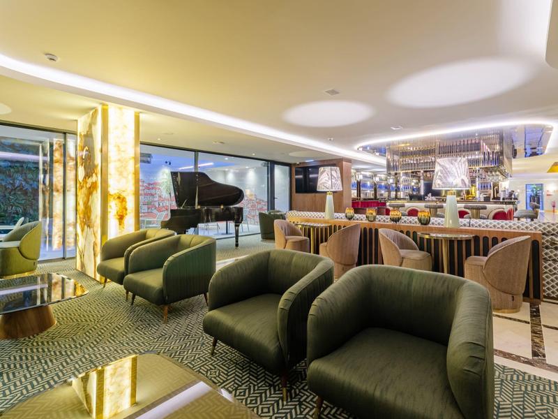 Lounge di un hotel moderno con poltrone verdi, bar e pianoforte in un ambiente elegante.