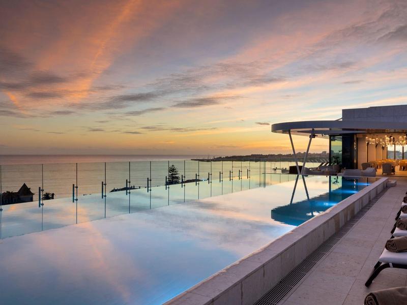 Basen z krawędzią bez krawędzi, szklanym ogrodzeniem i leżakami o zachodzie słońca nad morzem