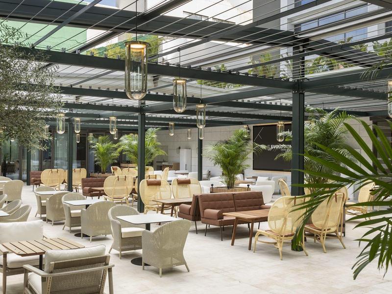 Nowoczesna restauracja hotelowa z eleganckimi miejscami do siedzenia i wieloma roślinami pod szklanym dachem.