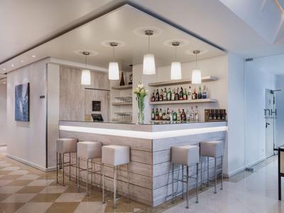 Moderner Hotelbar-Bereich mit Holztheke, Hockern und Hängelampen in heller Umgebung.