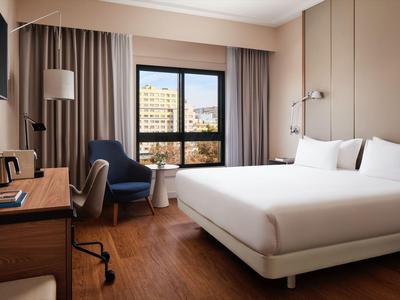 Modern eingerichtetes Hotelzimmer mit großem Bett, Fenster mit Stadtblick und Arbeitsplatz.