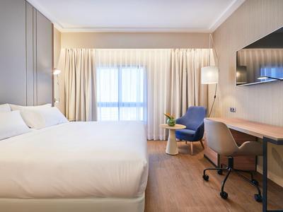 Modernes Hotelzimmer mit großem Bett, Schreibtisch, Fernseher und Stuhl vor einem Fenster mit Vorhängen.