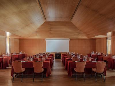 Ampia sala conferenze con pannelli di legno, file di tavoli e sedie, schermo di proiezione davanti.