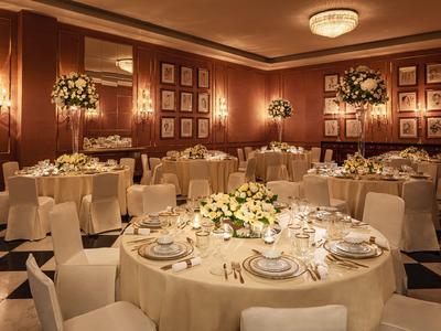 Sala da banchetto elegante con tavoli rotondi apparecchiati, sedie bianche e composizioni floreali.