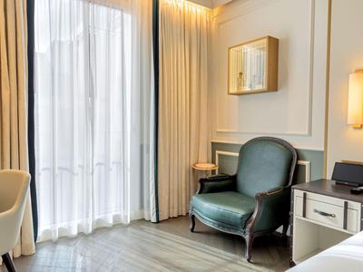 Camera d'hotel luminosa con poltrona in pelle verde, tende bianche e arredamento moderno.