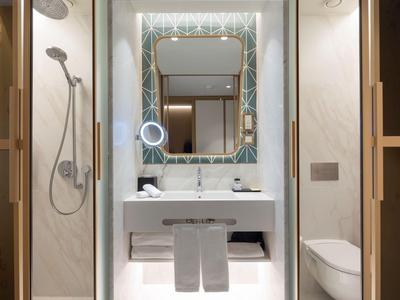 Bagno moderno in hotel con doccia a filo pavimento, specchio illuminato e piastrelle bianche.