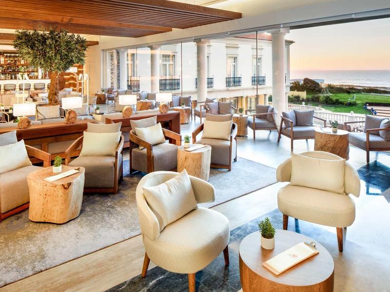 Area lounge luminosa con poltrone comode, tavoli in legno e vista sul giardino e sul mare.