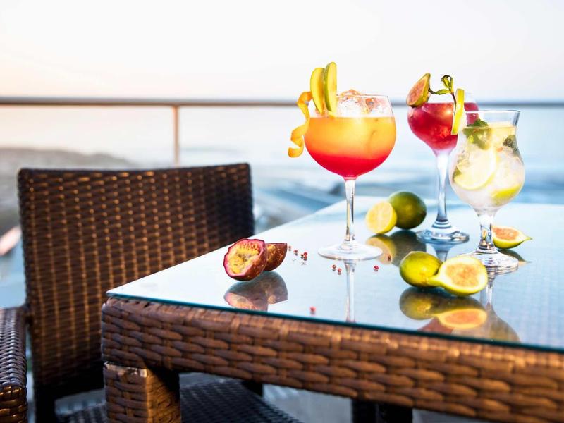 Tre cocktail colorati con frutta su un tavolo di vetro con vista mare e una sedia in vimini.