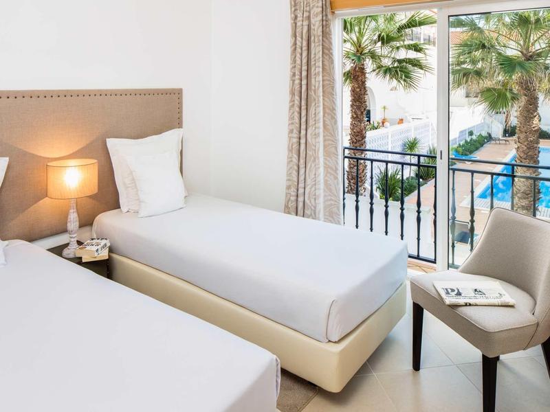 Camera d'hotel luminosa con due letti singoli, comodino, sedia e balcone con vista su palme e piscina.