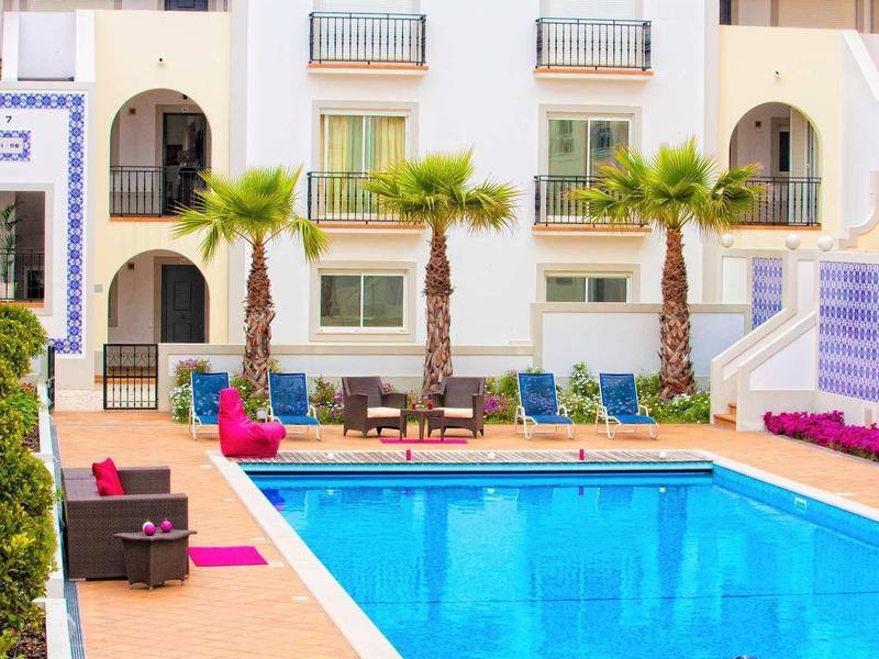 Area piscina invitante con lettini e palme davanti a un edificio alberghiero bianco.