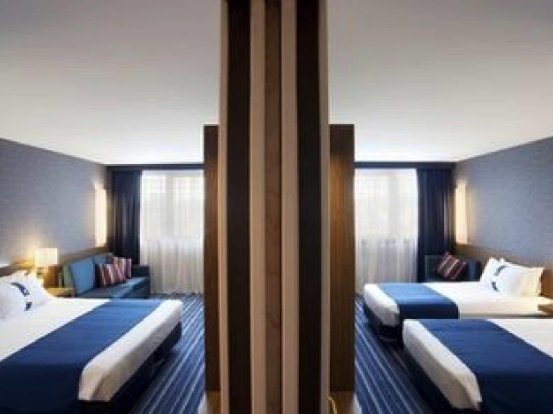 Camera d'hotel moderna con due letti, accenti blu e parete divisoria in uno spazio luminoso.