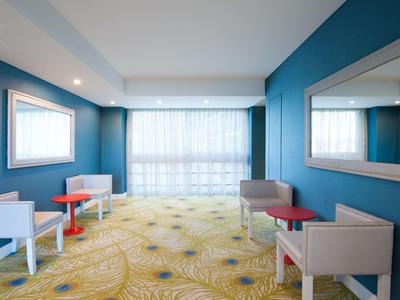 Flur mit blauen Wänden, gelbem Teppich, weißen Tischen, roten Stühlen und großen Spiegeln.