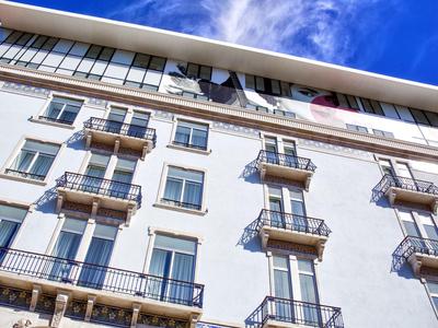 Fassadenansicht eines weißen Gebäudes mit kleinen Balkonen und Fenstern unter blauem Himmel.