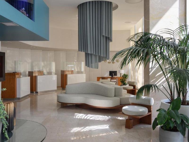 Lobby di hotel moderna con divano rotondo bianco, piante e grandi finestre che lasciano entrare la luce del giorno.