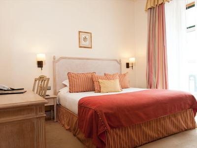 Camera d'hotel luminosa con letto matrimoniale, cuscini colorati e tende accanto a una scrivania.