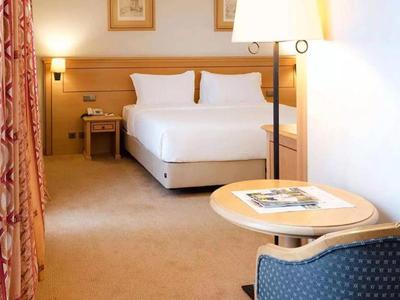 Camera d'hotel luminosa con letto matrimoniale, comodino, lampada da terra e piccolo tavolo con sedia.