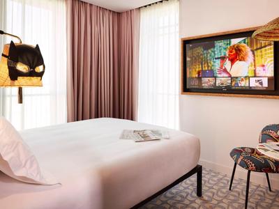 Camera d'hotel luminosa con letto grande, due sedie, tavolo rotondo e arte moderna alla parete.