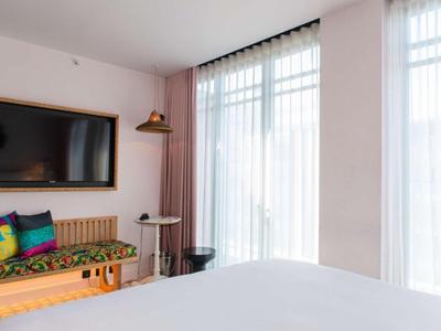 Lichte hotelkamer met groot bed, kleurrijke kussens, bank en groot raam met gordijnen.