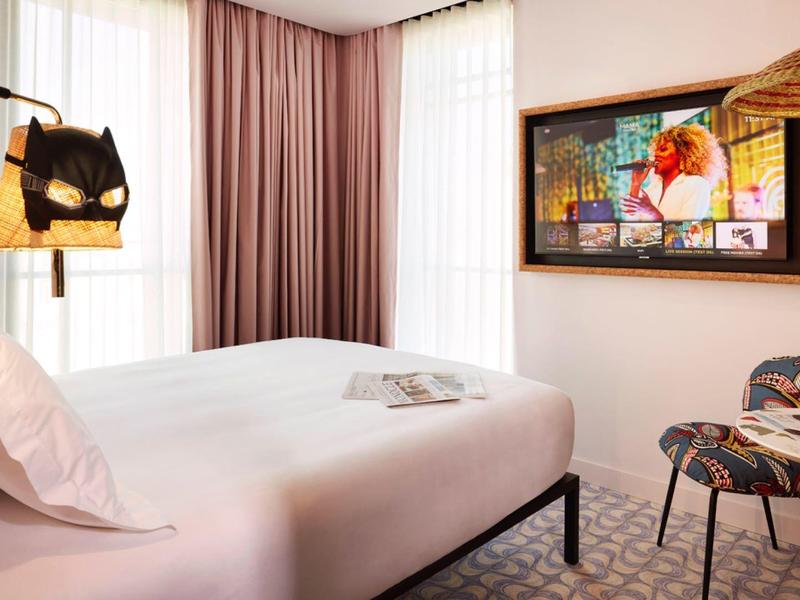 Camera d'hotel luminosa con letto grande, due sedie, tavolo rotondo e arte moderna alla parete.