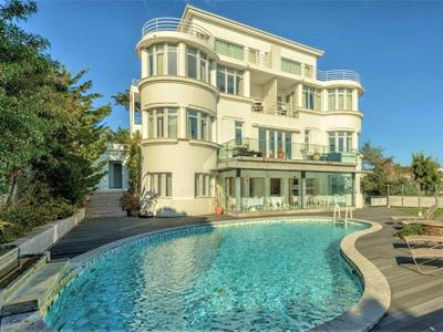 Großes helles Haus mit abgerundeten Balkonen, umgeben von Pool und Garten unter blauem Himmel.