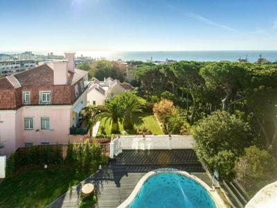 Vista di una piscina e alberi circostanti con un edificio rosa a sinistra e il mare sullo sfondo.