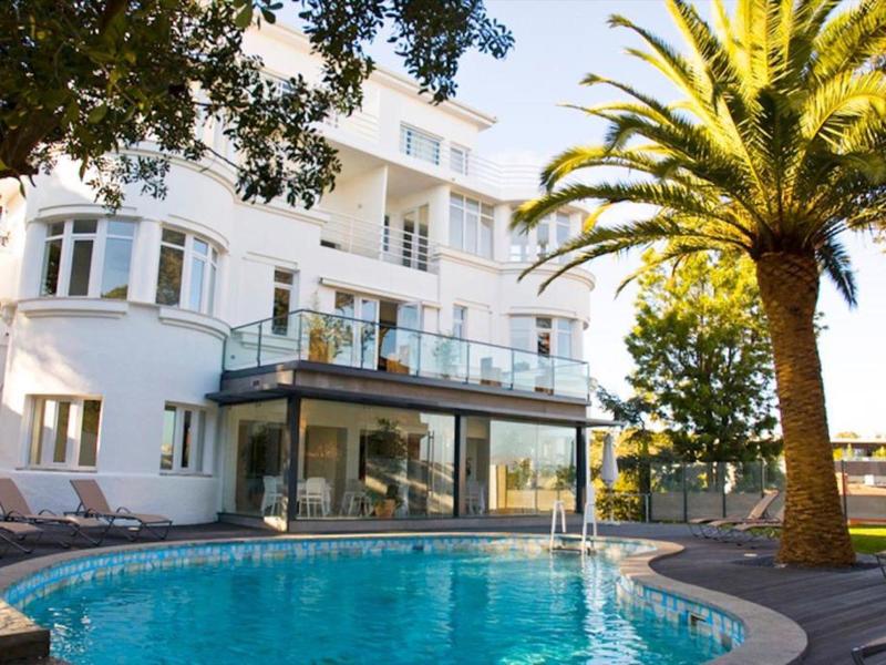 Großes weißes Haus mit Glasbalustrade und rundem blauen Pool, umgeben von Palmen und Bäumen.