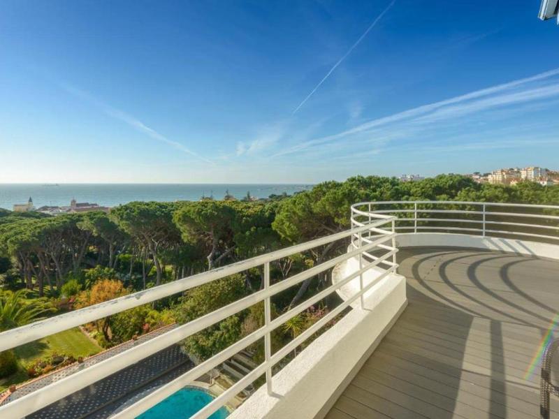 Großer Balkon mit weißem Geländer, Blick auf grüne Bäume und blauen Himmel am Meer.