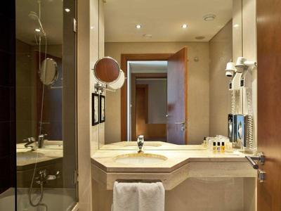 Helles Badezimmer mit weißem Waschbecken, Spiegel, Haartrockner und Glastür zur Dusche.