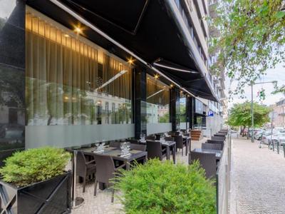 Modern outdoor restaurant terrace with tables, chairs, and green plants along a sidewalk.
