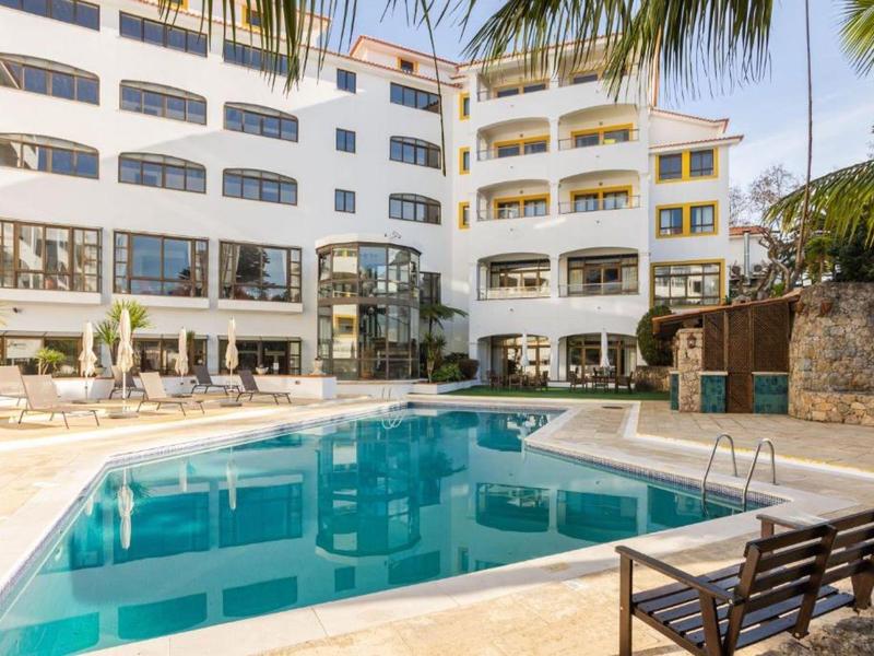 Hotel s venkovním bazénem, lehátky a palmami za slunečného počasí.