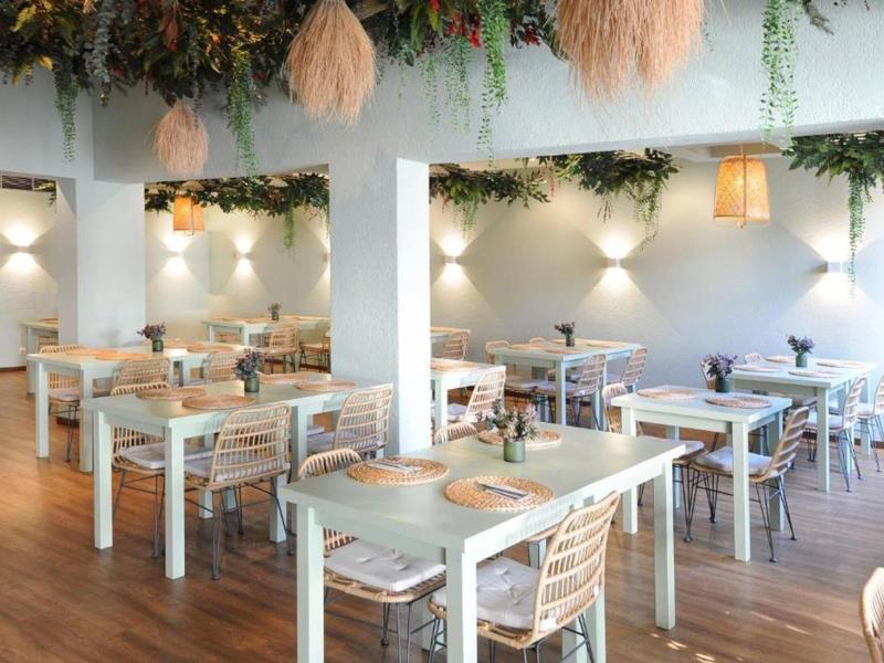 Světlá jídelna s bílými dřevěnými stoly, židlemi a zelenými dekoracemi na stropě.