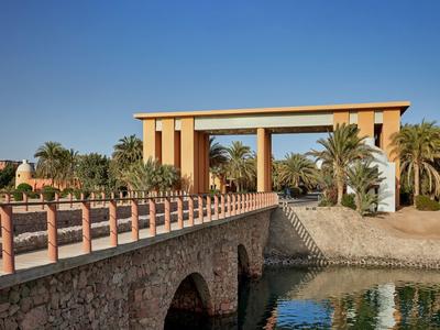 Puente de piedra sobre agua con portal marrón y palmeras bajo cielo despejado en zona hotelera.