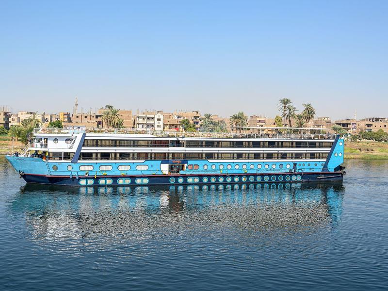 Barco de crucero largo en río tranquilo con edificios y palmeras al fondo.