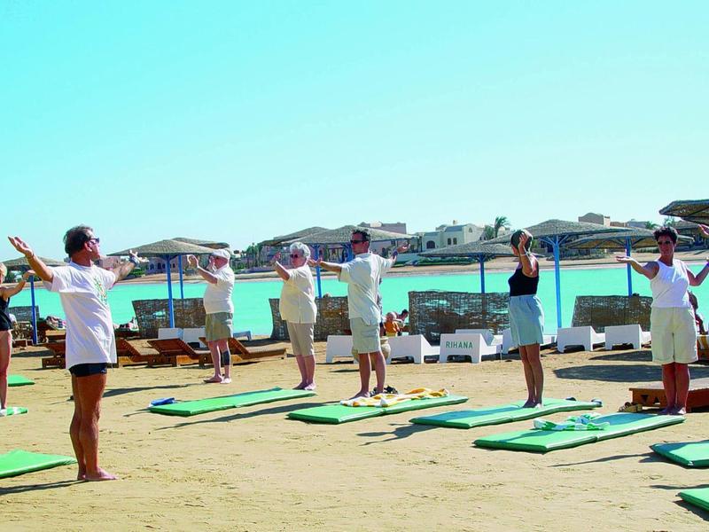 Personas hacen yoga en colchonetas verdes al aire libre con cielo azul y edificios al fondo.