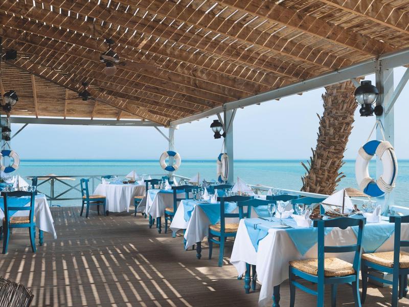Restaurante abierto frente al mar con techo de madera, mesas blancas y sillas azules, decoración marina.