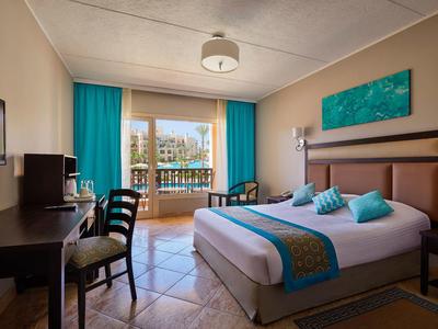 Hell eingerichtetes Hotelzimmer mit Doppelbett, Balkon, Schreibtisch und türkisfarbenen Vorhängen.