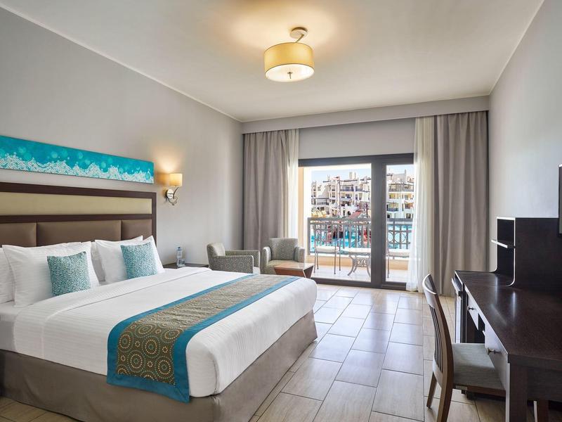 Modernes Hotelzimmer mit großem Bett, weißen Wänden, Balkon und Fernseher auf dunklem Holzschrank.