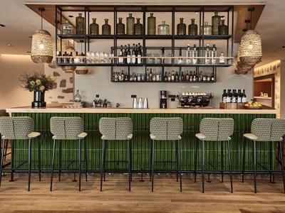 Moderner Hotelbarbereich mit grüner Theke, Barhockern und dekorativen Flaschenregalen.