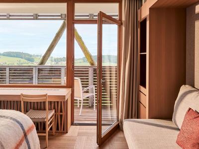 Gemütliches Zimmer mit Holzmöbeln, Bett, Sofa und offenem Balkon mit Blick auf grüne Hügel.