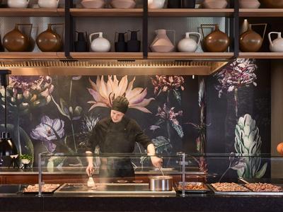 Koch bereitet Essen in moderner Küche mit floralem Hintergrund und Regalen voller Krüge vor.