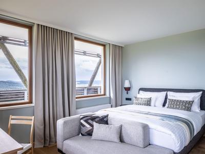 Modern eingerichtetes Schlafzimmer mit großem Bett, Sitzbank, Stuhl und Fenstern mit Blick nach draußen.