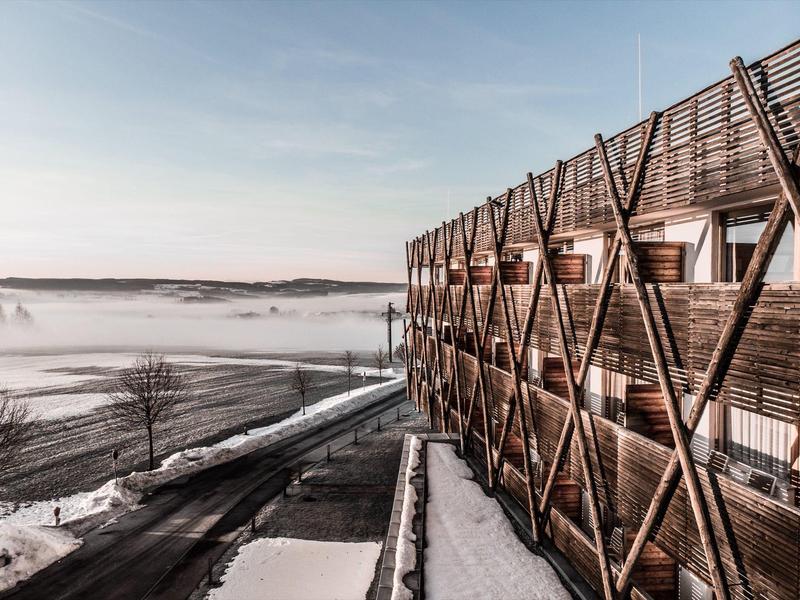 Modernes Holzhotel mit Balkonen neben einer schneebedeckten Straße und Nebel über dem Tal.