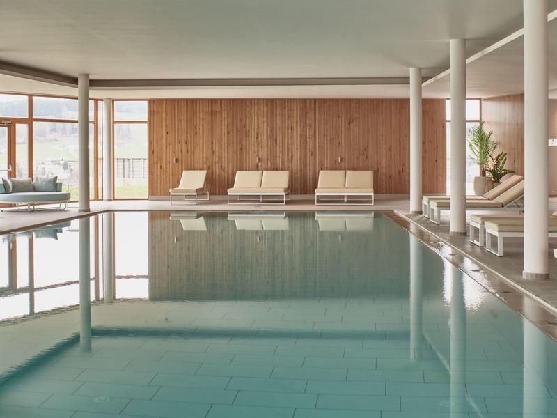 Innenbereich mit großem Schwimmbecken, Holzverkleidung und mehreren weißen Liegestühlen.