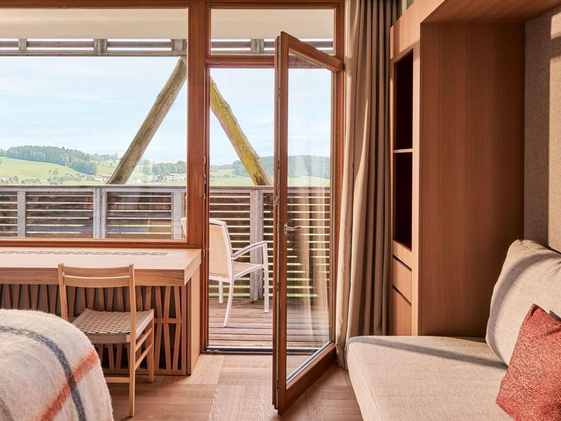 Gemütliches Zimmer mit Holzmöbeln, Bett, Sofa und offenem Balkon mit Blick auf grüne Hügel.