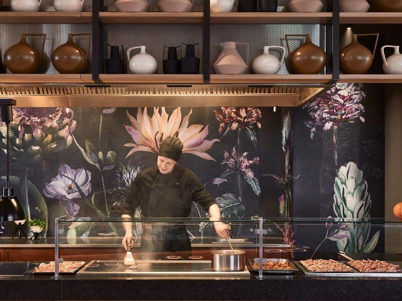 Koch in schwarzer Kleidung arbeitet in moderner Küche mit großem Blumenmuster an Wand.