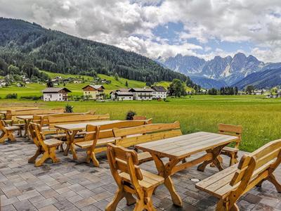 Freiluftbereich mit Holzbänken und Tischen, grünen Wiesen, Bergen und Häusern im Hintergrund.